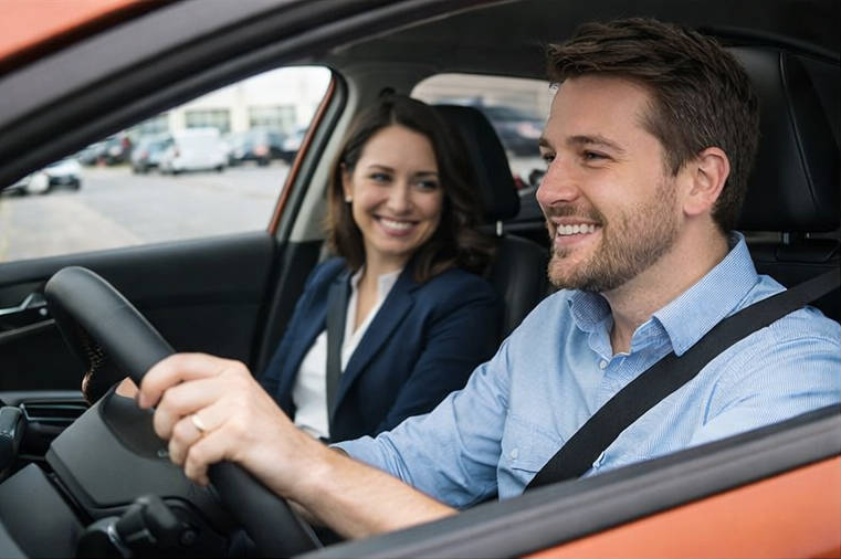 Experience a personalized test drive at Brossard Hyundai with a trusted expert Interior photo of an orange Hyundai Kona during a test drive in Brossard, showing a male client at the wheel and a Brossard Hyundai representative in the passenger seat, both smiling and buckled in.
